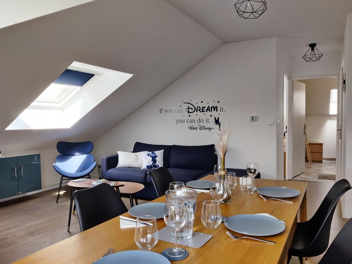 Séjour mansardé de l’appartement Akela avec canapé d’angle bleu marine et table à manger dressée sous les fenêtres de toit